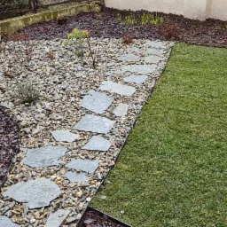 Świeżo założony trawnik z rolki graniczący z rabatą wysypaną kamieniami i ścieżką z nieregularnych płyt kamiennych, obrzeża wykończone czarnym bordem.