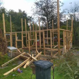 Szkielet drewnianej konstrukcji budynku w trakcie budowy, widoczne elementy pionowe i poziome, w tle zieleń drzew i krzewów, na pierwszym planie kosz na śmieci i resztki materiałów budowlanych.