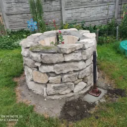 Studnia ogrodowa z kamienia polnego, z widocznym fragmentem instalacji pompy, otoczona trawnikiem i ogrodzeniem panelowym.