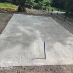 Wylany betonowy fundament pod budowę, widoczny niebieski pręt do poziomowania, ziemia wokół, w tle częściowo widoczne drzewa i ogrodzenie.