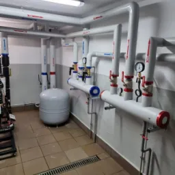 Węzeł cieplny z białymi rurami oznaczonymi strzałkami, manometry, zawory z czerwonymi pokrętłami oraz szary zbiornik wyrównawczy w pomieszczeniu z brązowymi płytkami podłogowymi i kratką ściekową.
