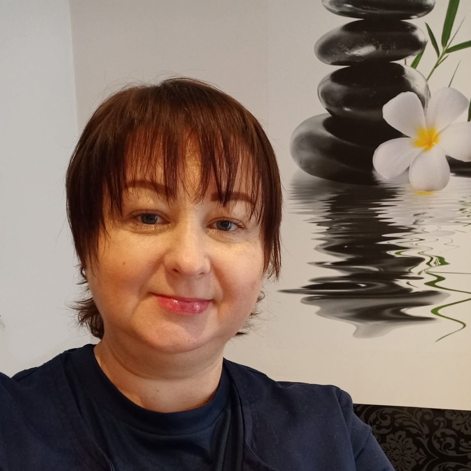 Portret kobiety z krótkimi brązowymi włosami i błyszczykiem na ustach, na tle dekoracji ze stosu kamieni i kwiatu w salonie.