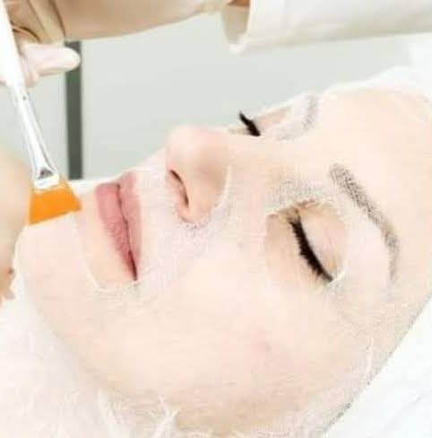 Nakładanie maski kosmetycznej pędzlem na twarz kobiety leżącej na białym podłożu. Zabieg pielęgnacyjny w zbliżeniu.