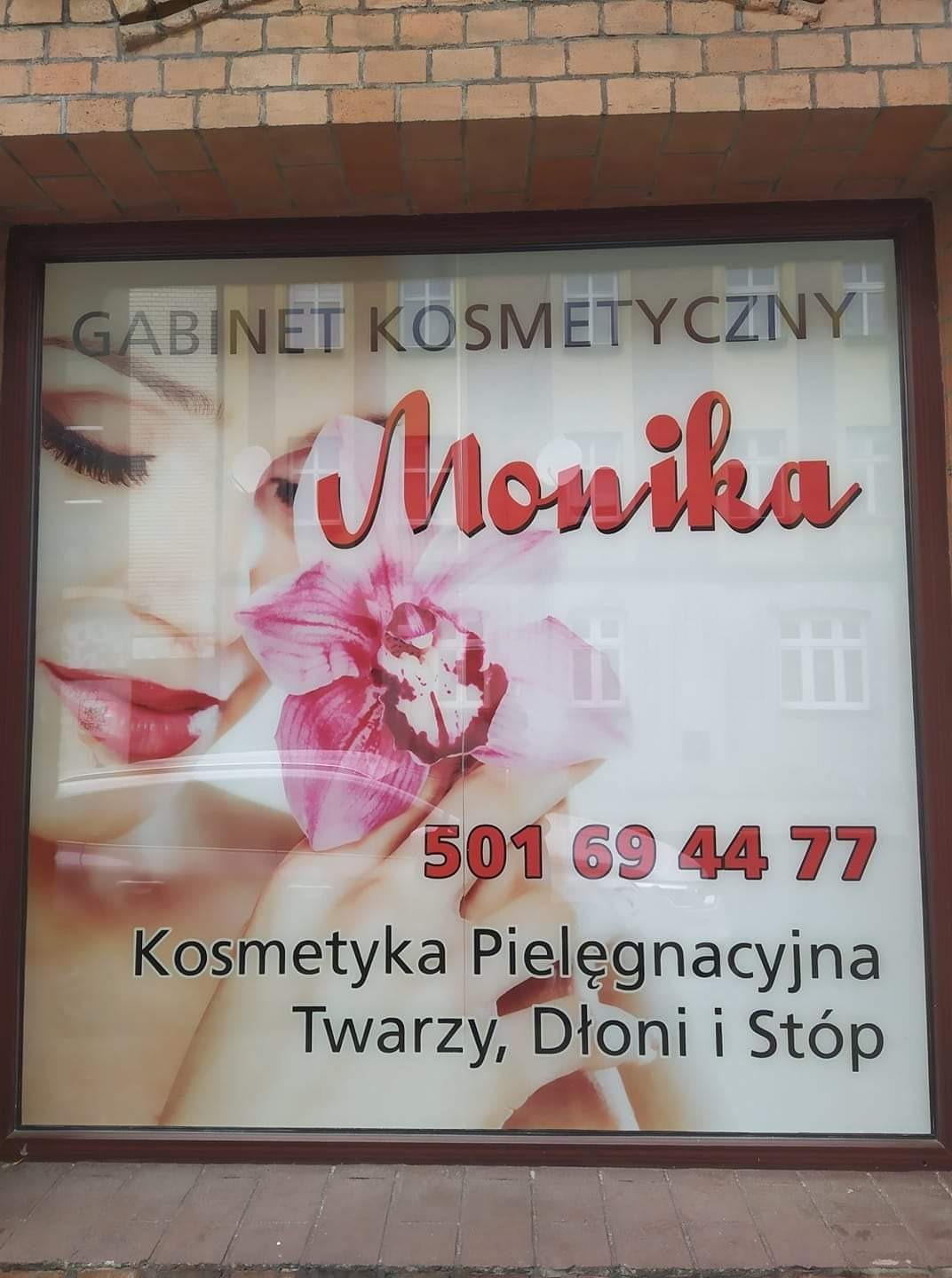 Szyld gabinetu kosmetycznego z wizerunkiem kobiety, numerem telefonu i informacją o kosmetyce pielęgnacyjnej twarzy, dłoni i stóp, umieszczony w oknie budynku.