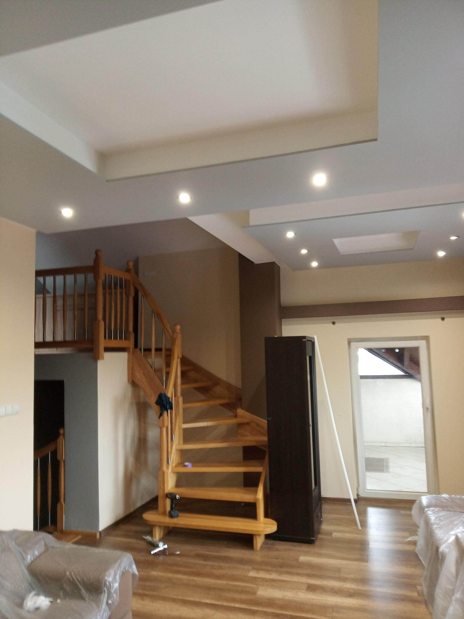 Wnętrze domu w trakcie prac wykończeniowych: drewniane schody z balustradą, częściowo zakryty mebel folią ochronną, widoczne narzędzia na podłodze, oświetlenie sufitowe.
