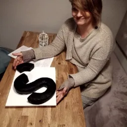 Uśmiechnięta kobieta prezentuje na drewnianym stole czarny, ręcznie wykonany element w kształcie litery S, obok leży jego fragment, całość na białej podkładce.