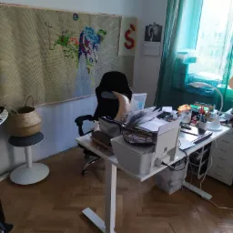 Rozgardiasz na biurku w pracowni artystycznej, z widocznym nieukończonym gobelinem na ścianie, koszem z włóczką i drukarką na blacie.