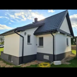 Dom jednorodzinny po renowacji elewacji, z jasną fasadą i ciemną podmurówką oraz dachem, widoczne rury spustowe i zwinięty wąż ogrodowy na trawniku.