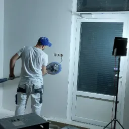 Pracownik w białych spodniach i niebieskiej czapce szlifuje ścianę szlifierką oscylacyjną, widoczne gniazdka elektryczne, lampa na statywie, odkurzacz przemysłowy i pojemniki na podłodze.