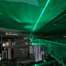 Laser krzyżowy emitujący zielone linie na surowym suficie podczas prac instalacyjnych, widoczne metalowe korytka kablowe i elementy konstrukcyjne budynku w tle.
