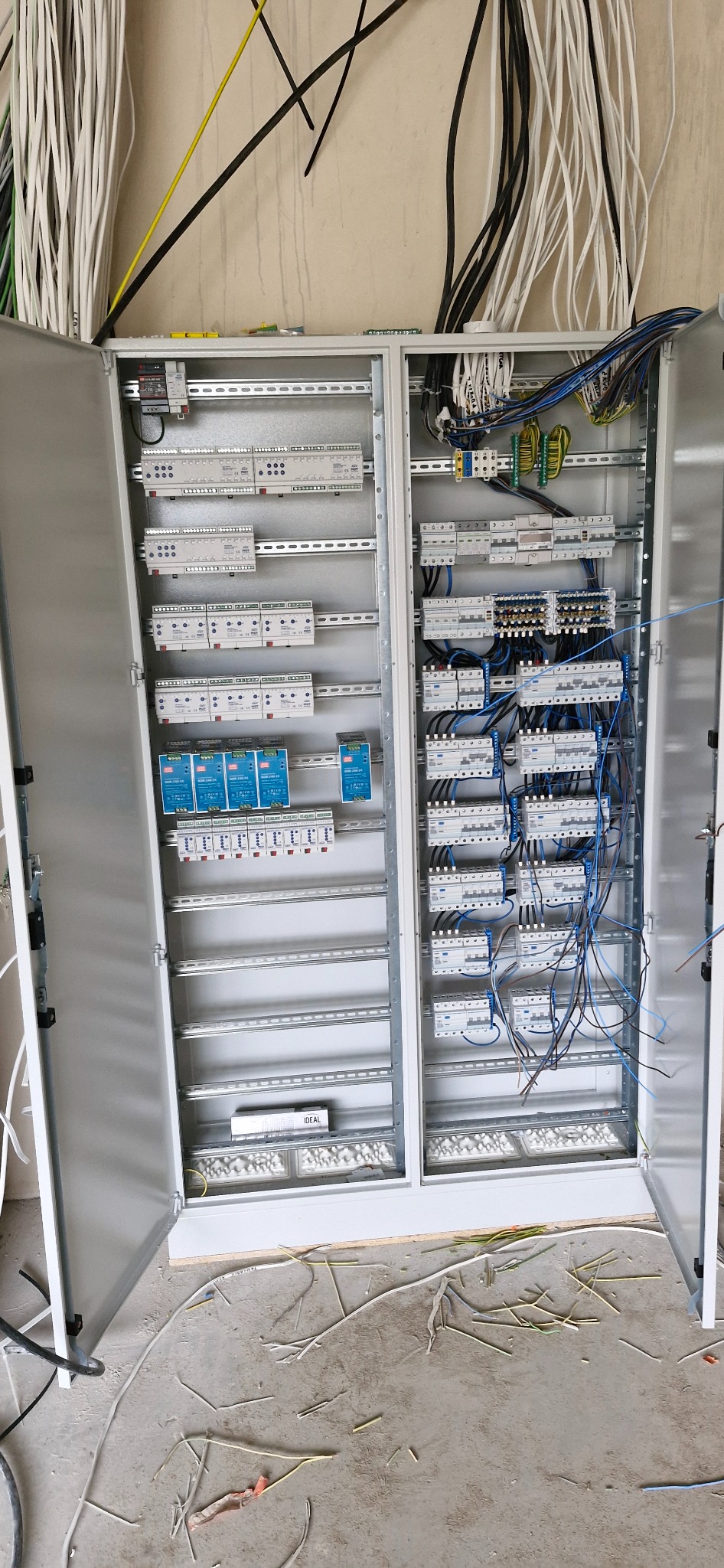 Otwarta szafa elektryczna z zainstalowanymi modułami, widoczne kable i przewody, na podłodze resztki kabli i fragment opakowania.