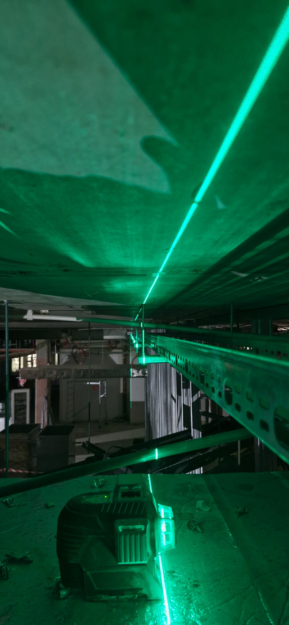 Laser krzyżowy emitujący zielone linie na surowym suficie podczas prac instalacyjnych, widoczne metalowe korytka kablowe i elementy konstrukcyjne budynku w tle.