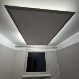 Szare podwieszane oświetlenie sufitowe w kształcie trapezu z ukrytym oświetleniem LED, widok na okno z odbiciem osoby, grzejnik i rury.