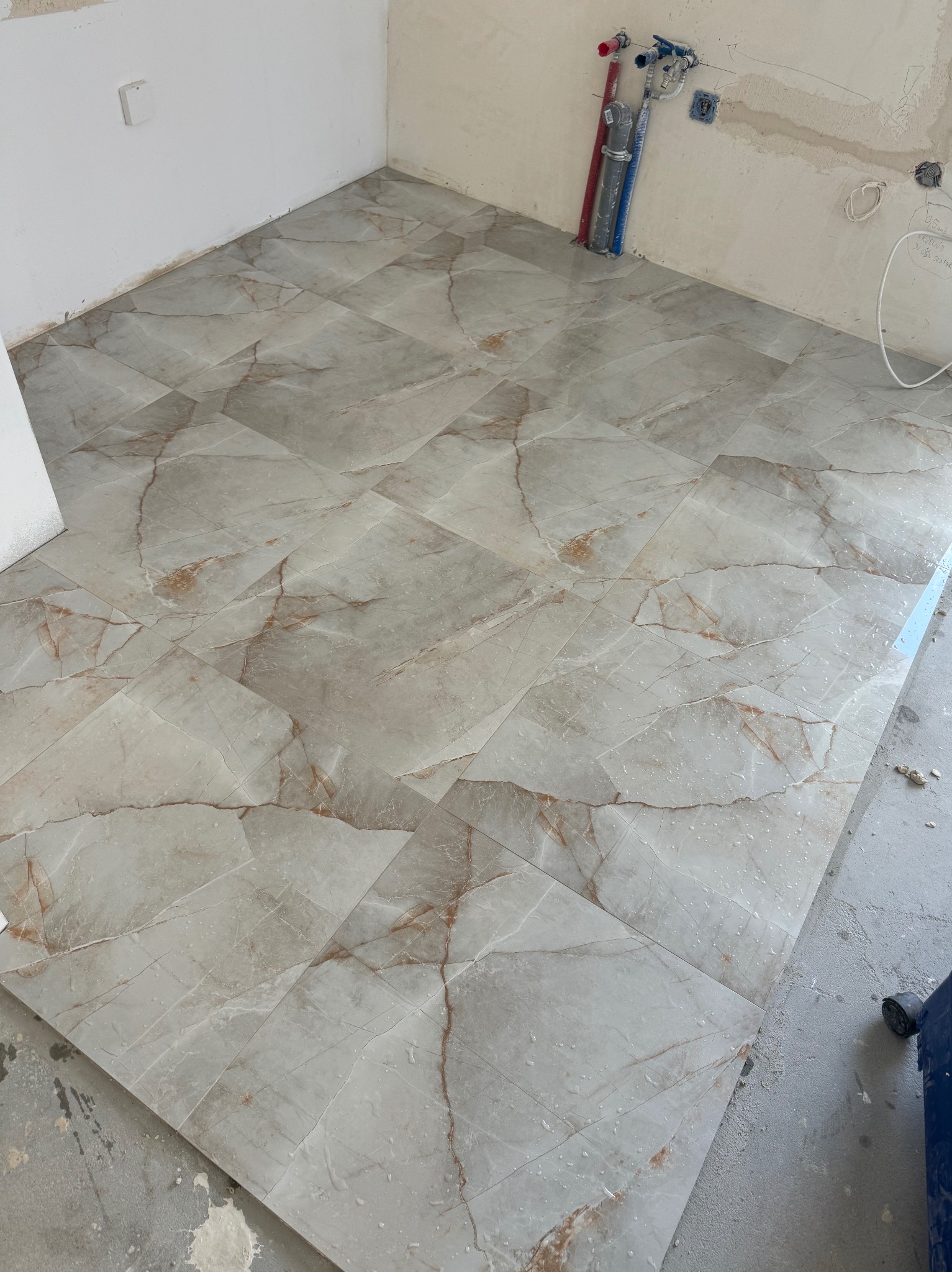 Ułożone płytki podłogowe imitujące marmur w trakcie remontu łazienki, widoczne przyłącza wodne na ścianie i pozostałości po pracach budowlanych.