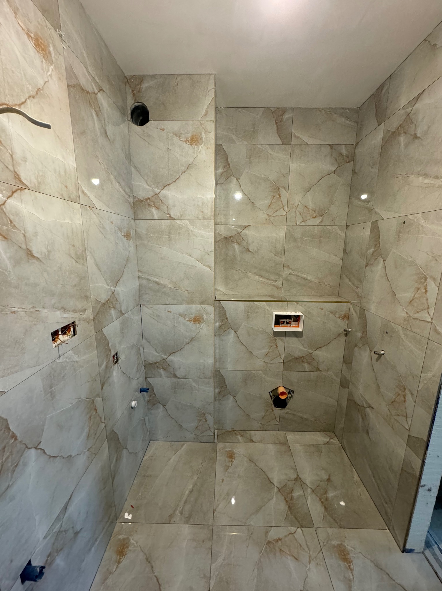 Wnętrze łazienki w trakcie prac wykończeniowych, widoczne płytki imitujące marmur na ścianach i podłodze, otwory na instalacje wodno-kanalizacyjne i elektryczne, brak armatury.