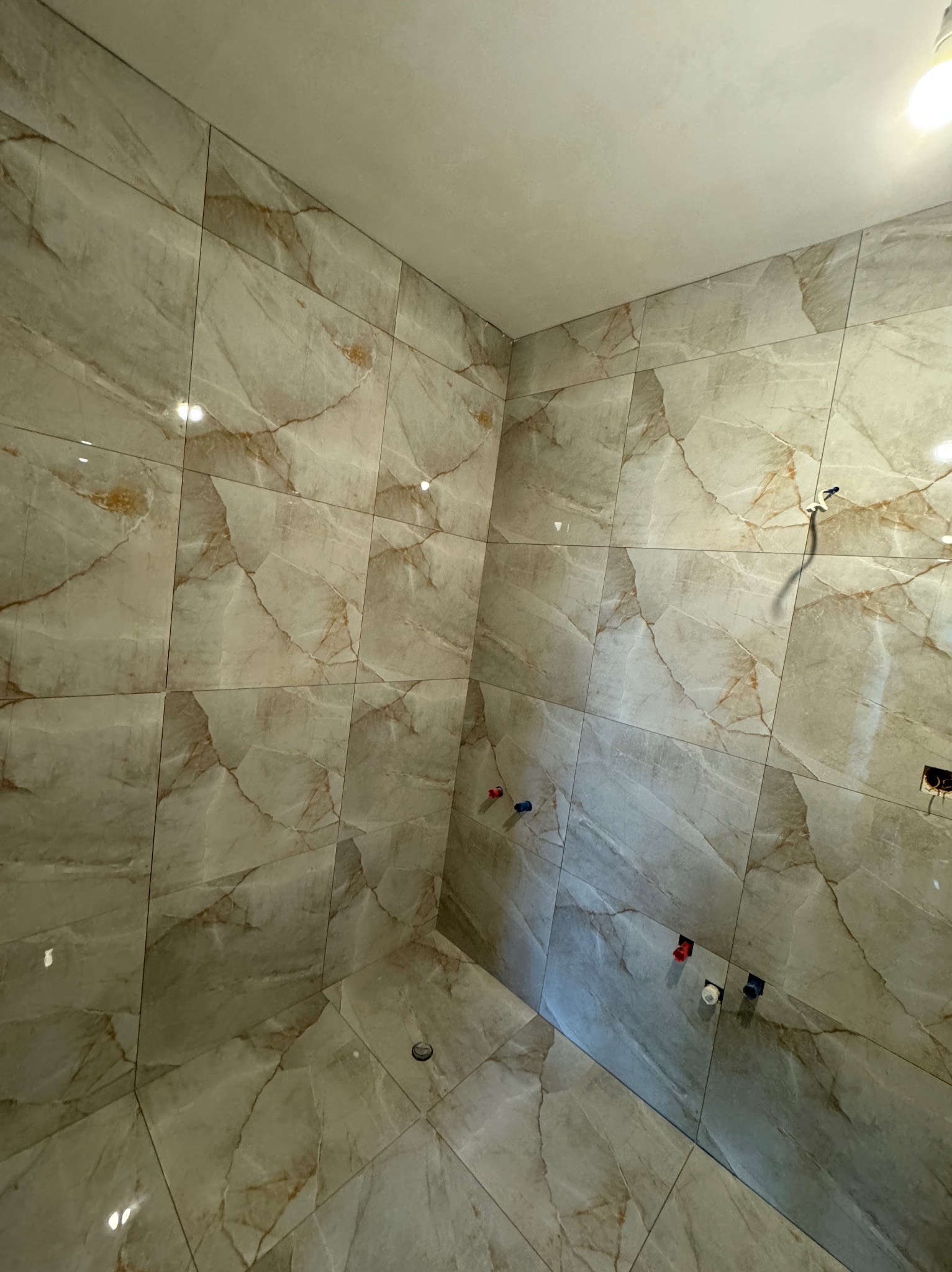 Wnętrze łazienki w trakcie prac wykończeniowych, widoczne płytki imitujące marmur na ścianach i podłodze, wystające rury z zaślepkami, surowy sufit i punktowe oświetlenie.