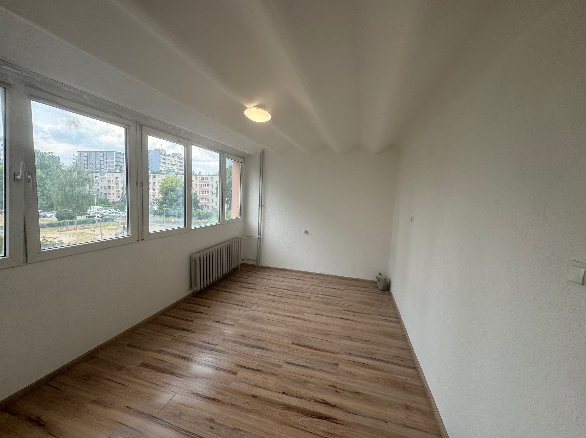 Pusty pokój po remoncie z panelami podłogowymi w odcieniach brązu, widok na osiedle przez duże okno, białe ściany i sufit z centralnym oświetleniem.