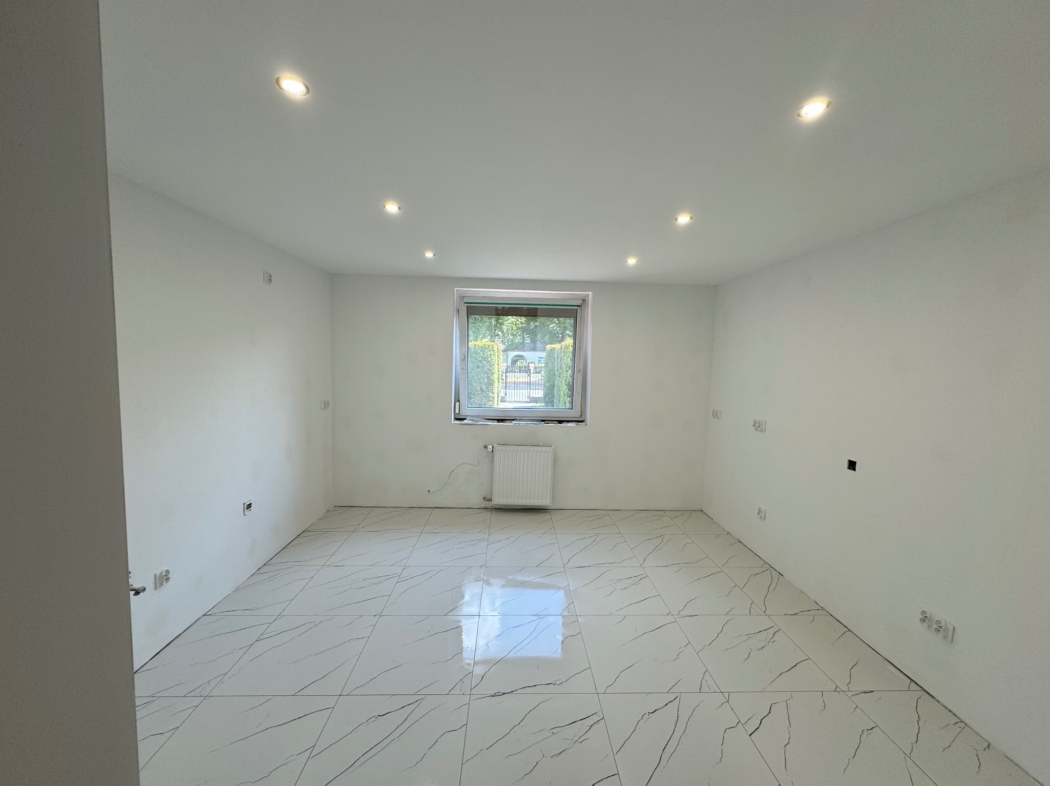 Puste, jasne pomieszczenie po remoncie z białymi ścianami, marmurową podłogą, wpuszczonymi lampami sufitowymi i grzejnikiem pod oknem z widokiem na zieleń.