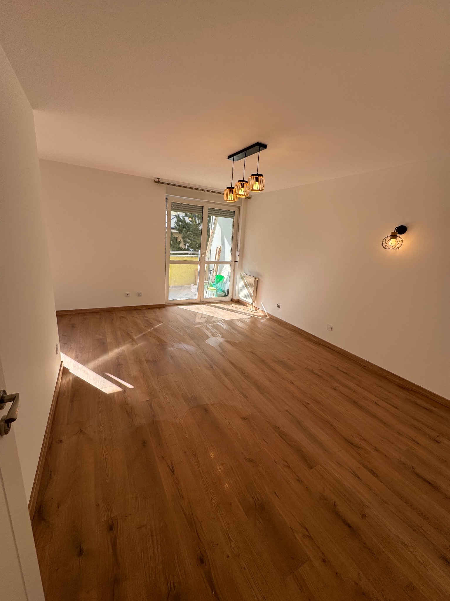 Jasne, puste pomieszczenie z drewnianą podłogą, oświetlone wiszącą lampą i kinkietem, z balkonem w tle.