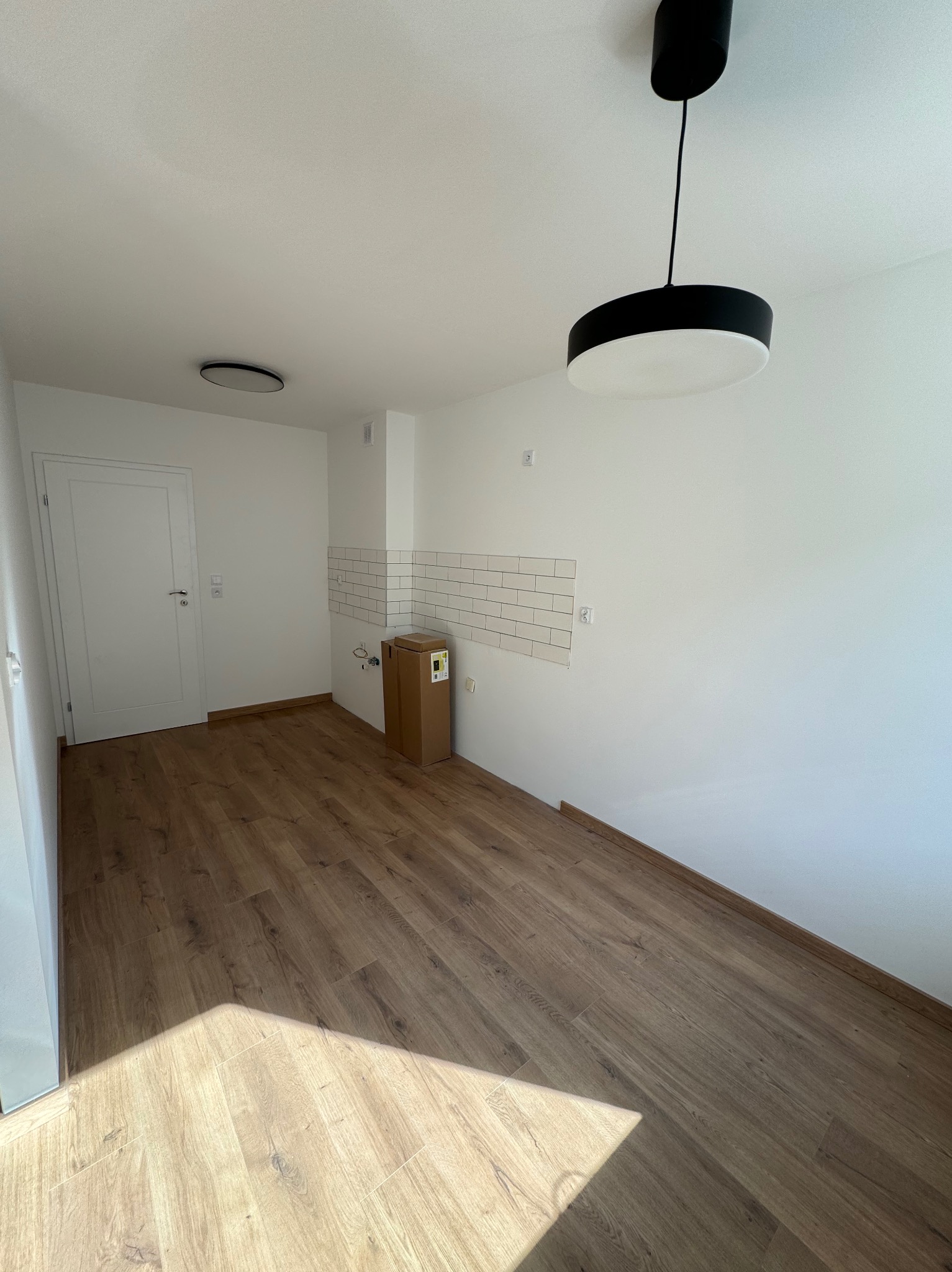 Puste pomieszczenie z panelami podłogowymi w odcieniach brązu, białe ściany z fragmentem ułożonych białych płytek w stylu cegiełki, kartonowe pudełko z wyposażeniem i nowoczesna lampa sufitowa.