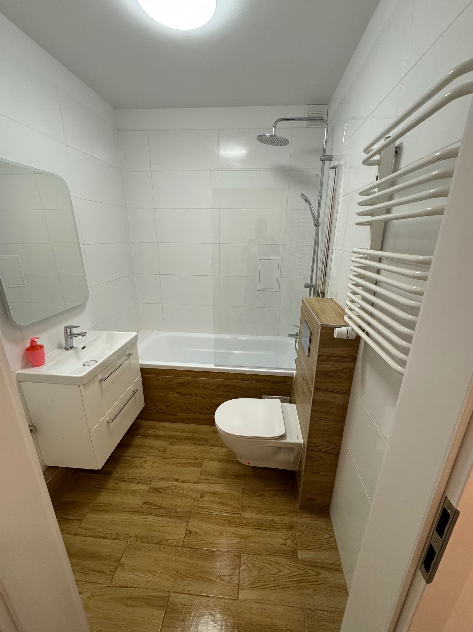 Nowoczesna, jasna łazienka z drewnopodobną podłogą, białymi płytkami ściennymi, wanną z parawanem, umywalką z szafką i toaletą typu Geberit.