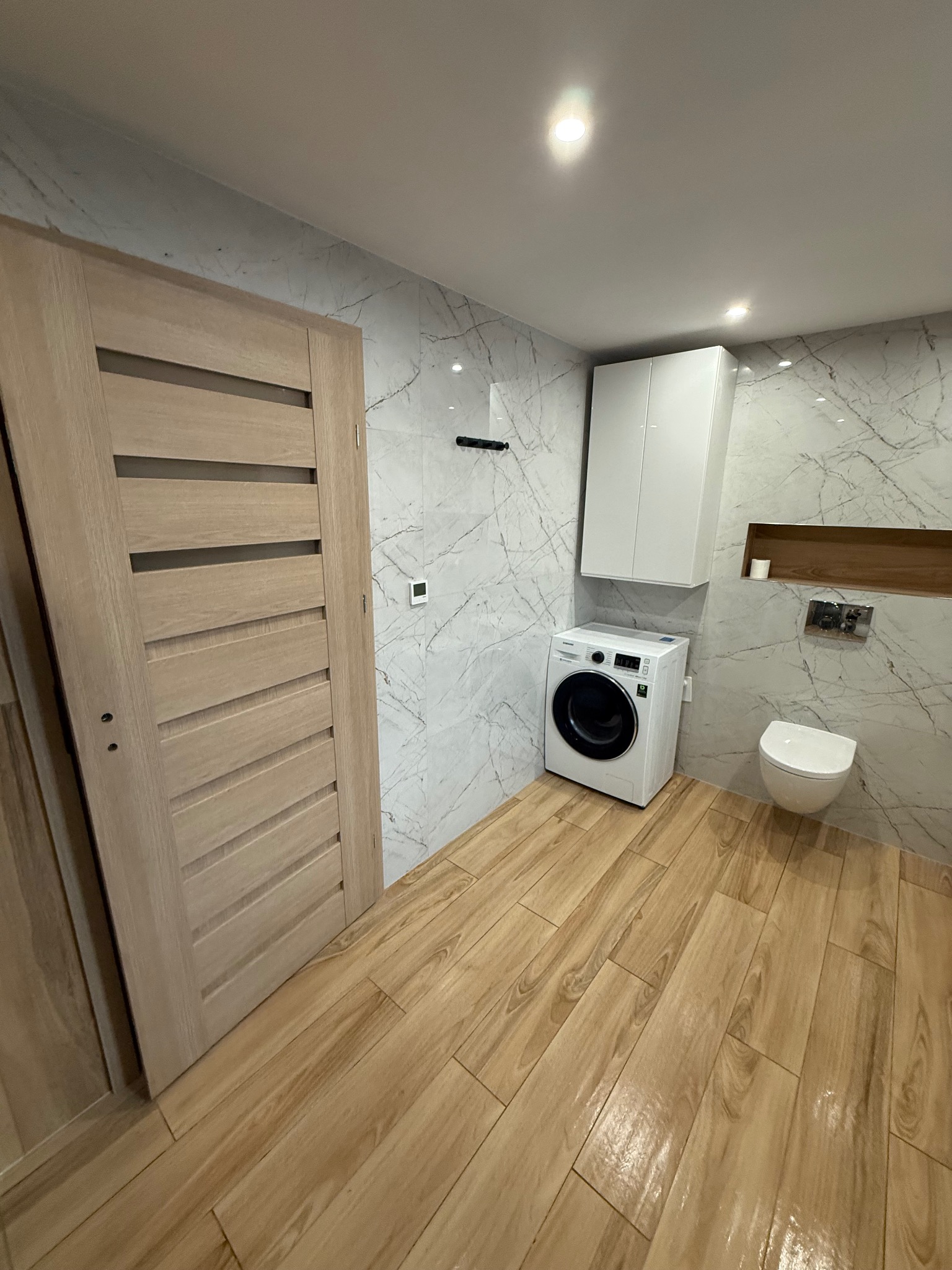 Nowoczesna łazienka z drewnopodobną podłogą, marmurowymi ścianami, białą pralką i podwieszaną toaletą. Jasne oświetlenie, minimalistyczny design.