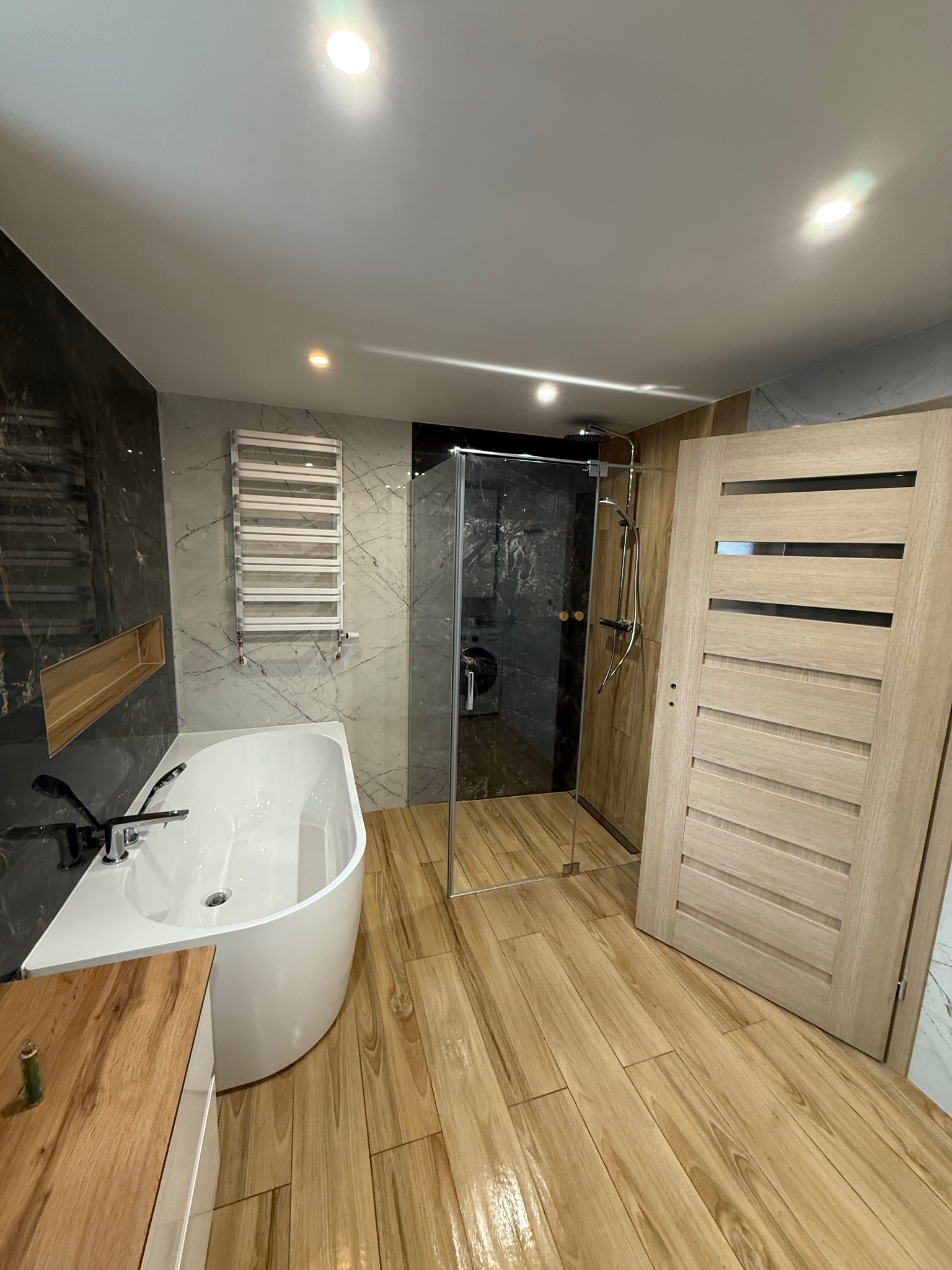 Nowoczesna łazienka z wanną, kabiną prysznicową i drewnianymi akcentami. Marmurowe płytki na ścianach, drewnopodobna podłoga, grzejnik drabinkowy.