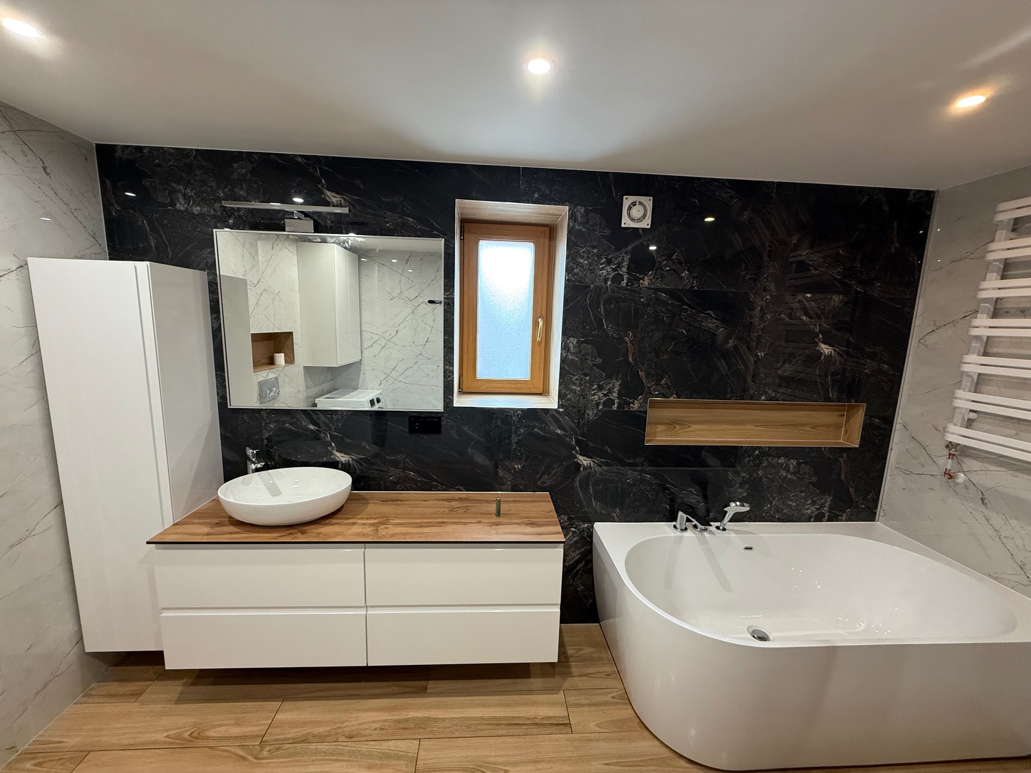 Nowoczesna łazienka z czarnymi marmurowymi płytkami, drewnianym blatem pod umywalką i białą wanną. Widoczne lustro i okno z drewnianą ramą.