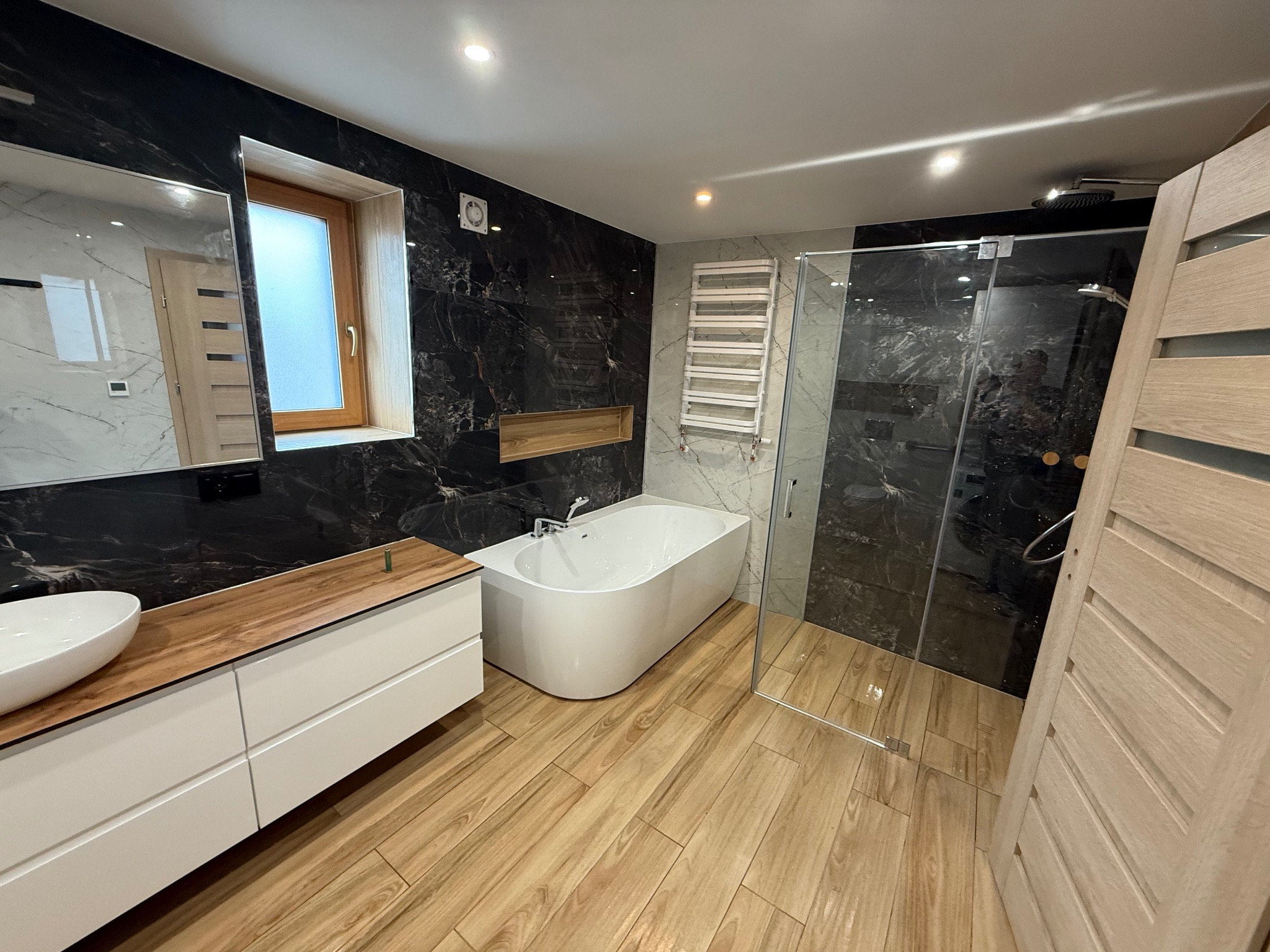 Nowoczesna łazienka z czarnymi marmurowymi płytkami, drewnianą podłogą i białymi meblami. Kabina prysznicowa ze szklanymi drzwiami i grzejnik drabinkowy.