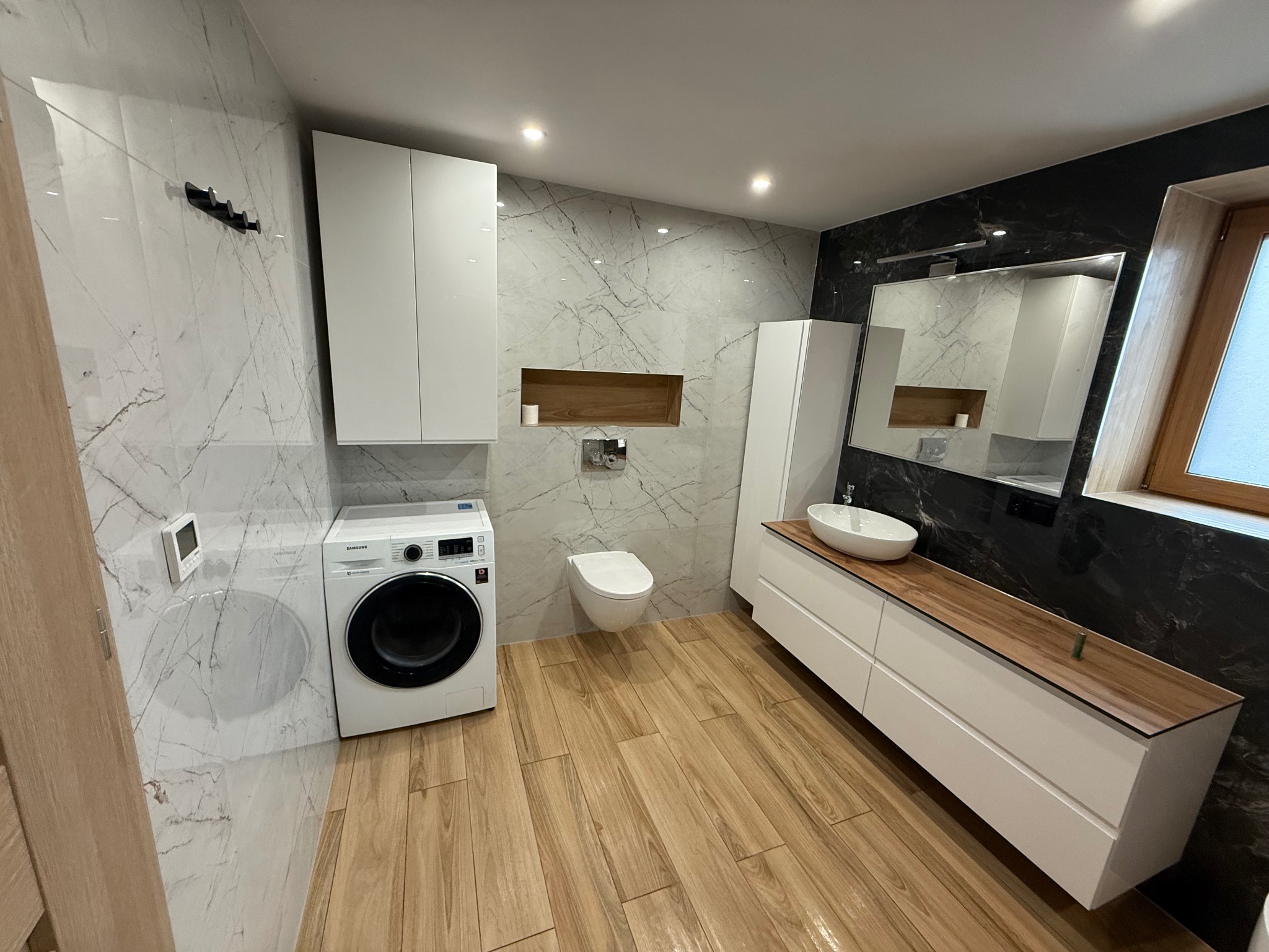 Nowoczesna łazienka z marmurowymi płytkami, drewnianą podłogą i białymi meblami. Pralka Samsung wkomponowana w zabudowę. Minimalistyczny design z akcentami drewna.