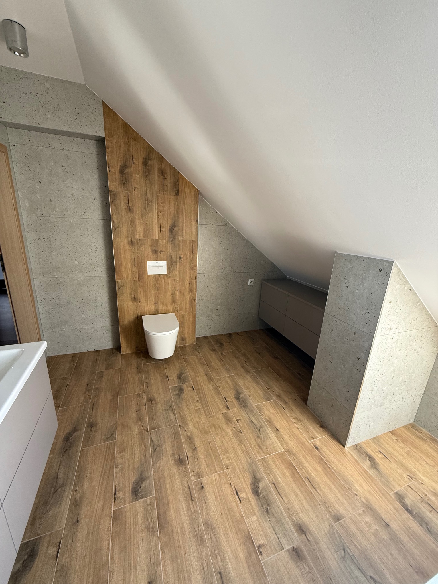 Nowoczesna łazienka na poddaszu z drewnopodobną podłogą i szarą okładziną ścienną. Uwagę przykuwa toaleta pod skosem i zabudowa z szufladami.