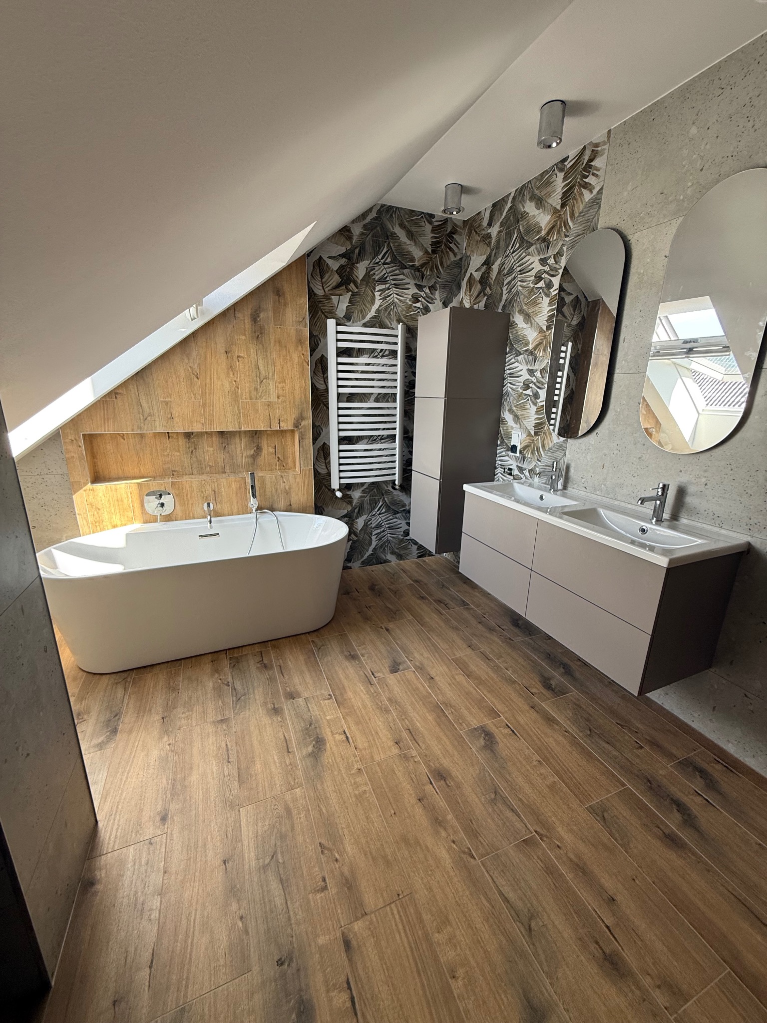 Nowoczesna łazienka na poddaszu z drewnianą podłogą, wolnostojącą wanną i umywalką z szafką. Ściany wykończone płytkami imitującymi drewno i kamień.