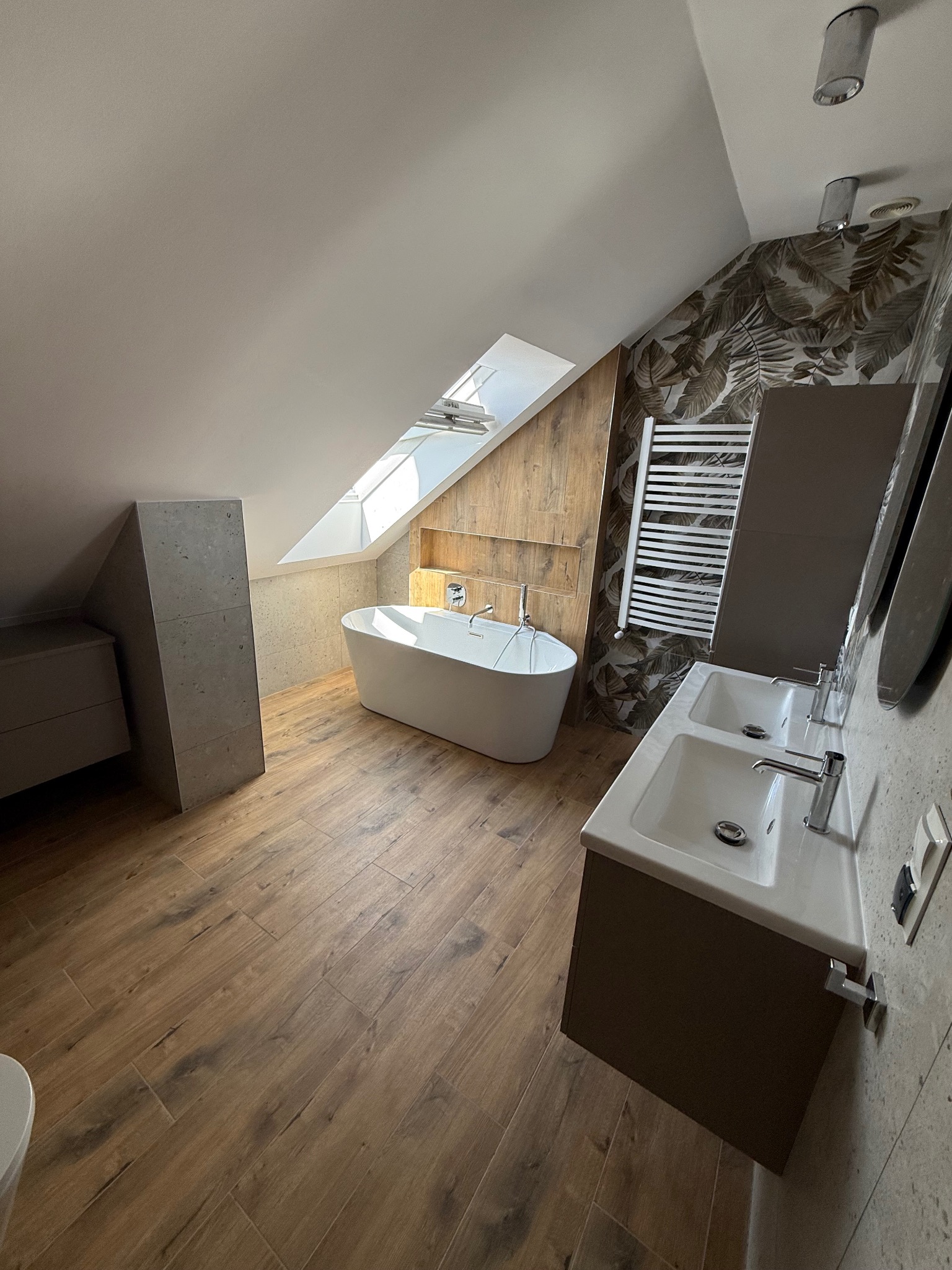 Nowoczesna łazienka na poddaszu z wolnostojącą wanną, dwoma umywalkami i grzejnikiem drabinkowym. Drewnopodobne płytki i akcenty roślinne na ścianie.