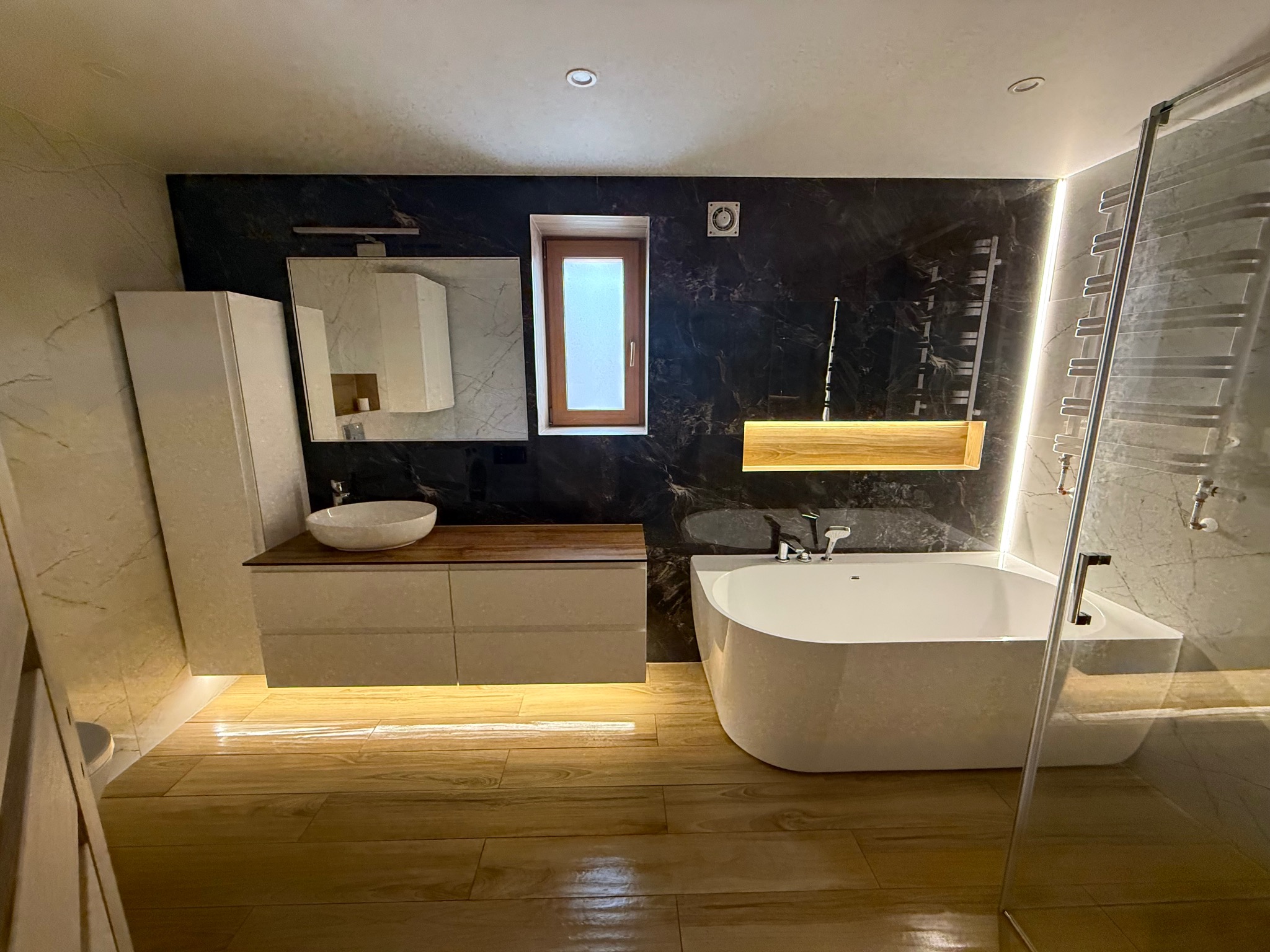 Nowoczesna łazienka z podświetlaną wanną, drewnianą podłogą i czarnymi marmurowymi ścianami. Umywalka nablatowa i szafka z oświetleniem LED. Okno w drewnianej ramie.
