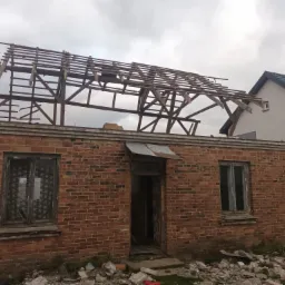 Częściowo zdemontowana więźba dachowa na ceglanym budynku z widocznymi starymi oknami i gruzem wokół, obok nowy dom.