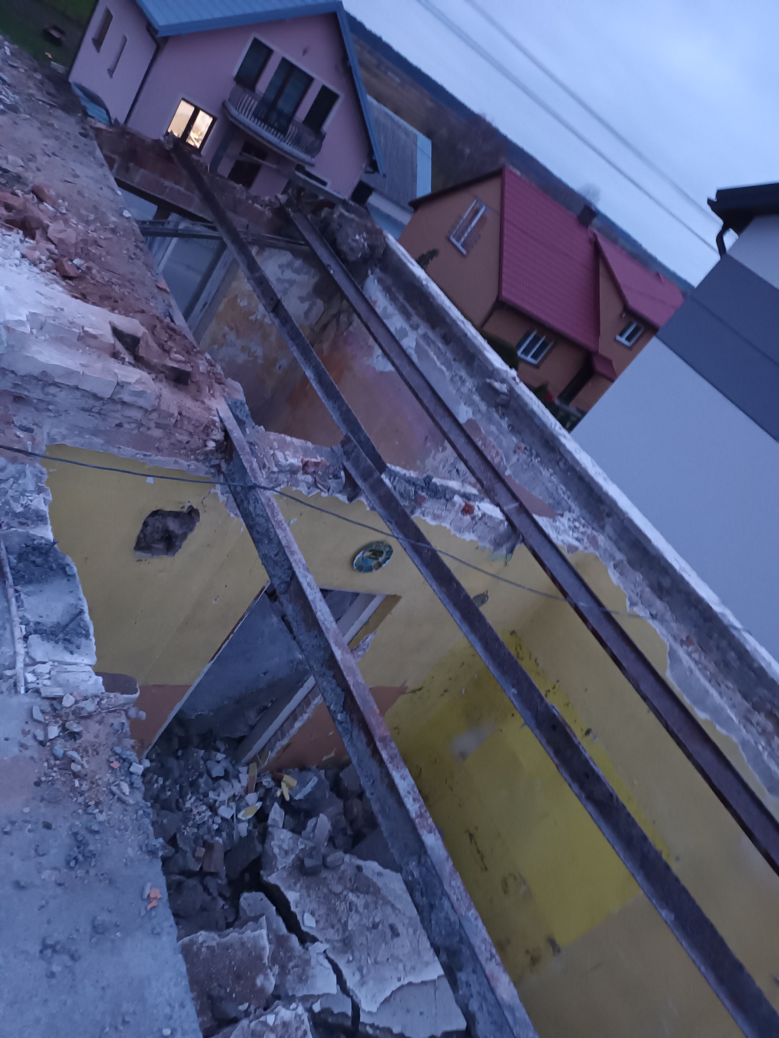 Rozbiórka stropu z widocznymi elementami konstrukcyjnymi i gruzem, żółta ściana w tle, ujęcie z góry na tle zabudowań i pochmurnego nieba.