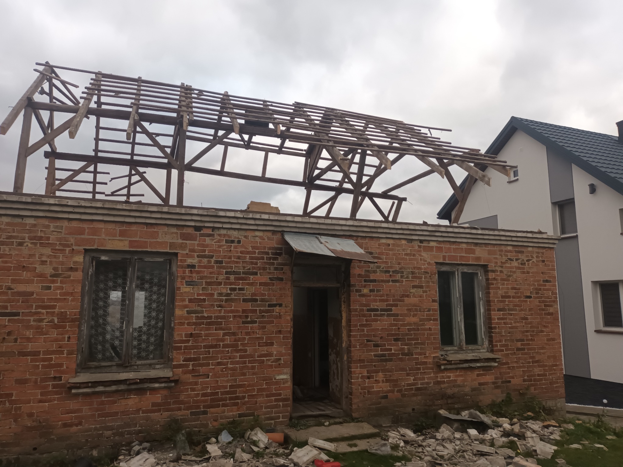 Częściowo zdemontowana więźba dachowa na ceglanym budynku z widocznymi starymi oknami i gruzem wokół, obok nowy dom.