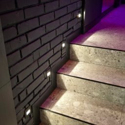 Instalacje Elektryczne TOMASZ ŁYSZKOWSKI - Stylowe schody z terazzo, podświetlone kwadratowymi lampkami w murze z cegły pomalowanej na szaro, oświetlone fioletowym światłem z góry.