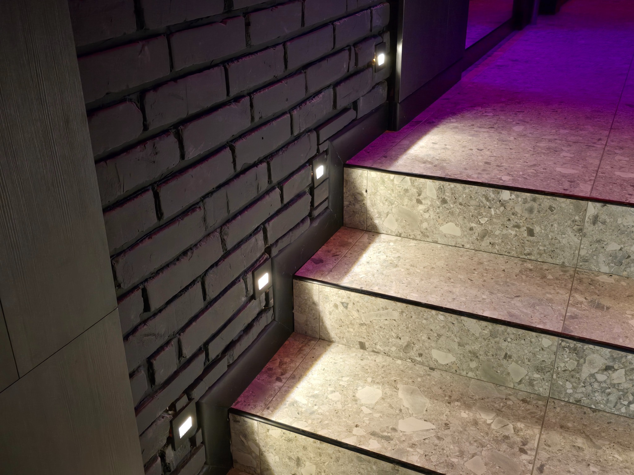 Stylowe schody z terazzo, podświetlone kwadratowymi lampkami w murze z cegły pomalowanej na szaro, oświetlone fioletowym światłem z góry.