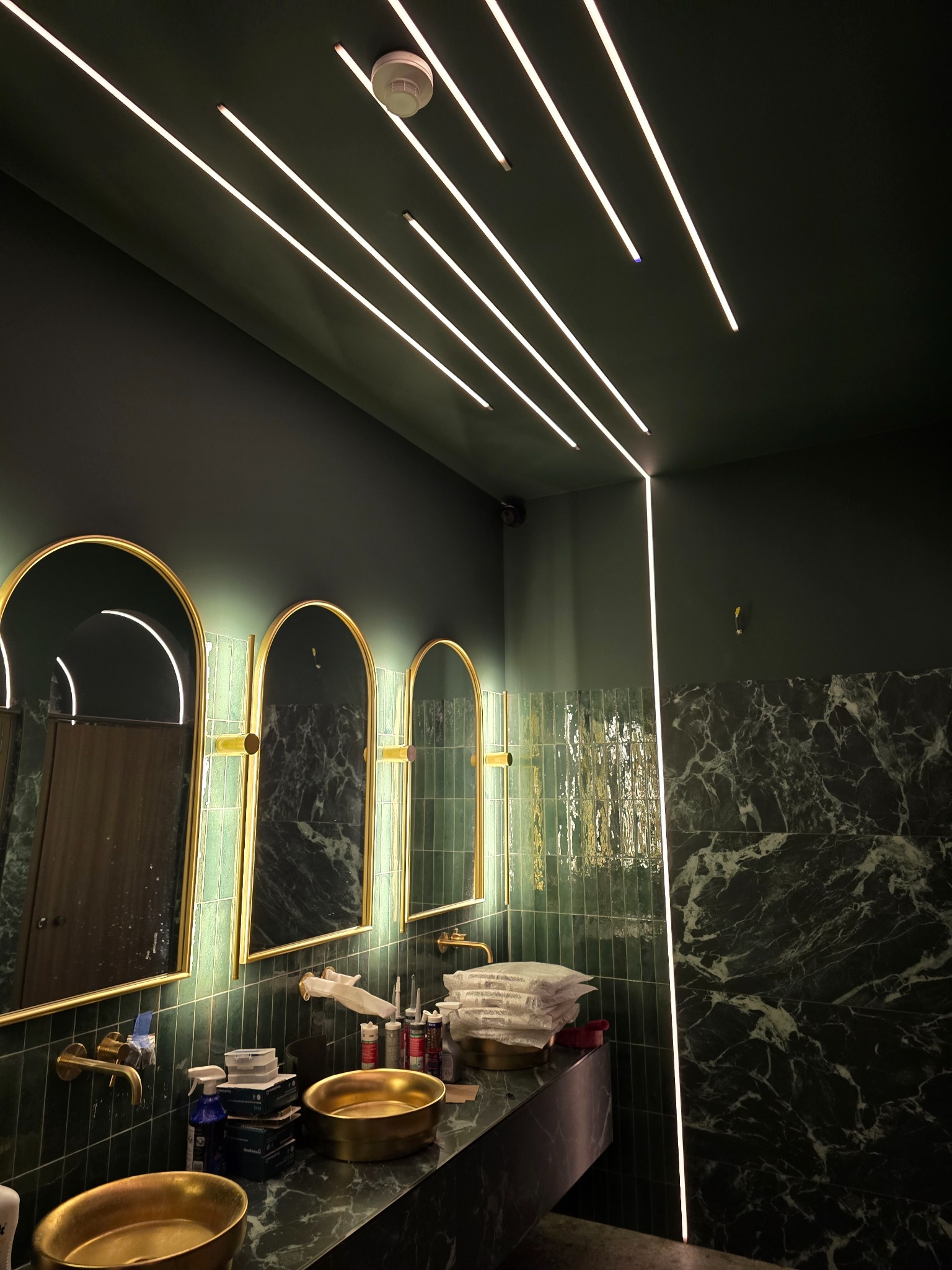 Nowoczesna łazienka w odcieniach zieleni z marmurowymi płytkami, złotymi umywalkami i podświetlanymi lustrami oraz liniowym oświetleniem LED na suficie.