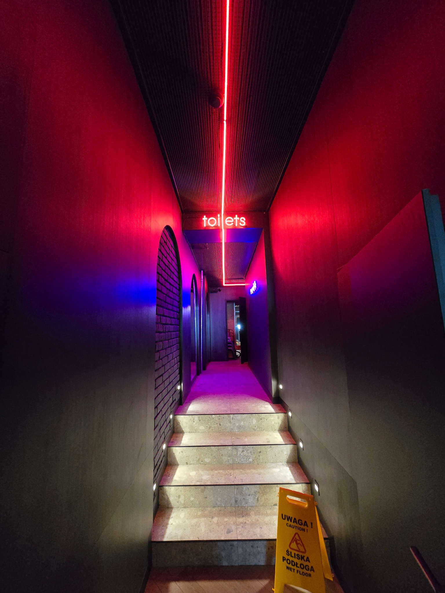 Nowoczesne oświetlenie schodów prowadzących do toalet, z neonowym napisem i ostrzegawczym znakiem 'Śliska podłoga'. Ciemne ściany i subtelne światła tworzą unikalny klimat.