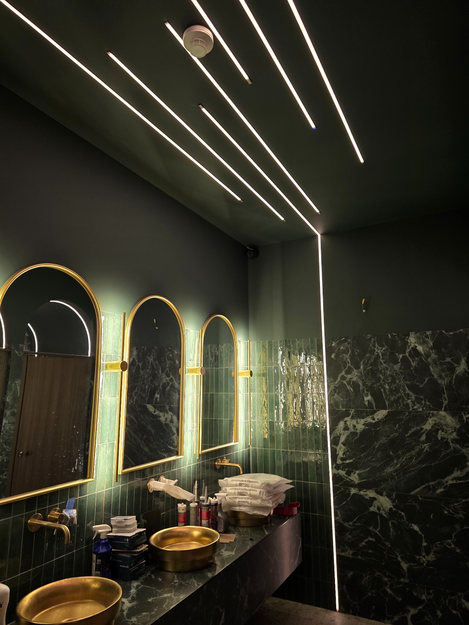Nowoczesna łazienka w odcieniach zieleni z marmurowymi płytkami, złotymi umywalkami i lustrami oraz liniowym oświetleniem LED na suficie. Widoczne akcesoria łazienkowe.