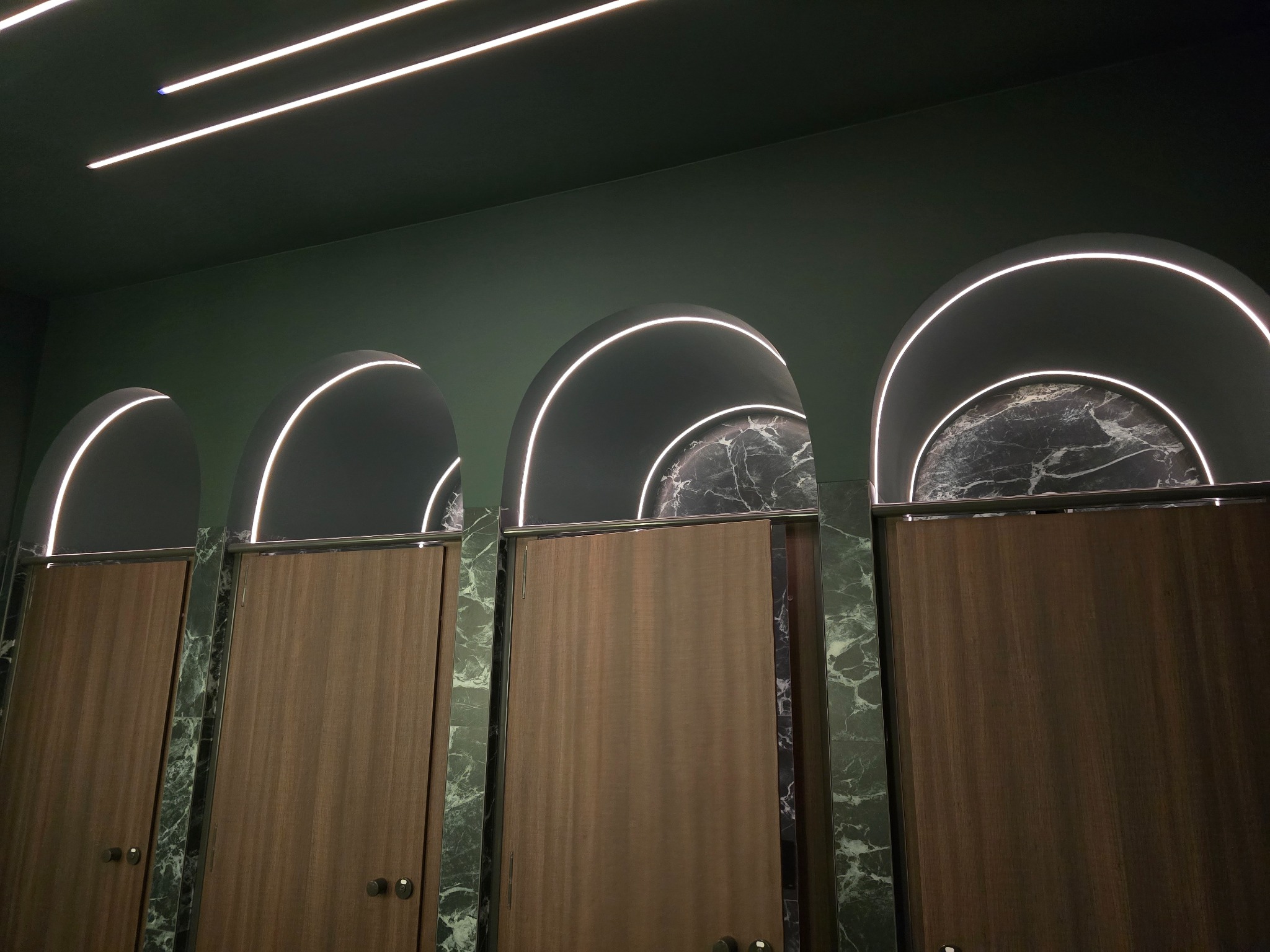 Toaleta publiczna z kabinami oświetlonymi łukami LED i marmurowymi ścianami. Nowoczesny design z zielonymi ścianami i drewnianymi drzwiami.