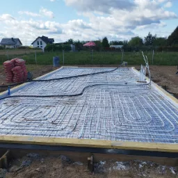 Przygotowanie do wylewki pod ogrzewanie podłogowe: widoczne rury grzewcze rozłożone na siatce metalowej, obramowanie z desek, w tle worki z zaprawą, domy i błękitne niebo z chmurami.