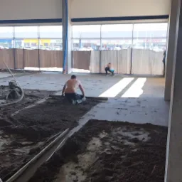 Dwóch mężczyzn wyrównuje mokrą zaprawę na dużej powierzchni wewnątrz budynku z oknami, widoczne narzędzia i elementy konstrukcyjne hali.