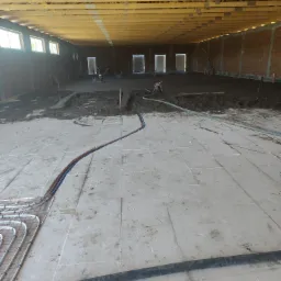 Układanie ogrzewania podłogowego w dużej hali. Część podłogi z wylewką, część z instalacją grzewczą. Widoczne narzędzia i ekipa w tle. Surowy stan wykończenia.