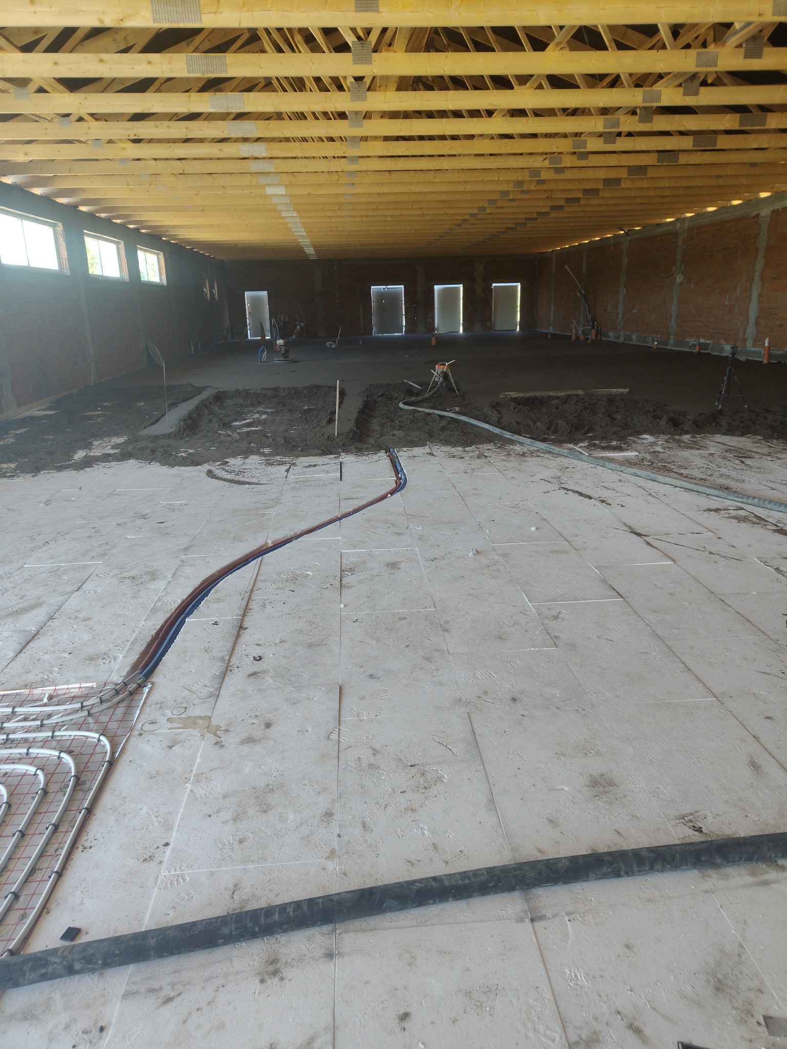 Układanie ogrzewania podłogowego w dużej hali. Część podłogi z wylewką, część z instalacją grzewczą. Widoczne narzędzia i ekipa w tle. Surowy stan wykończenia.