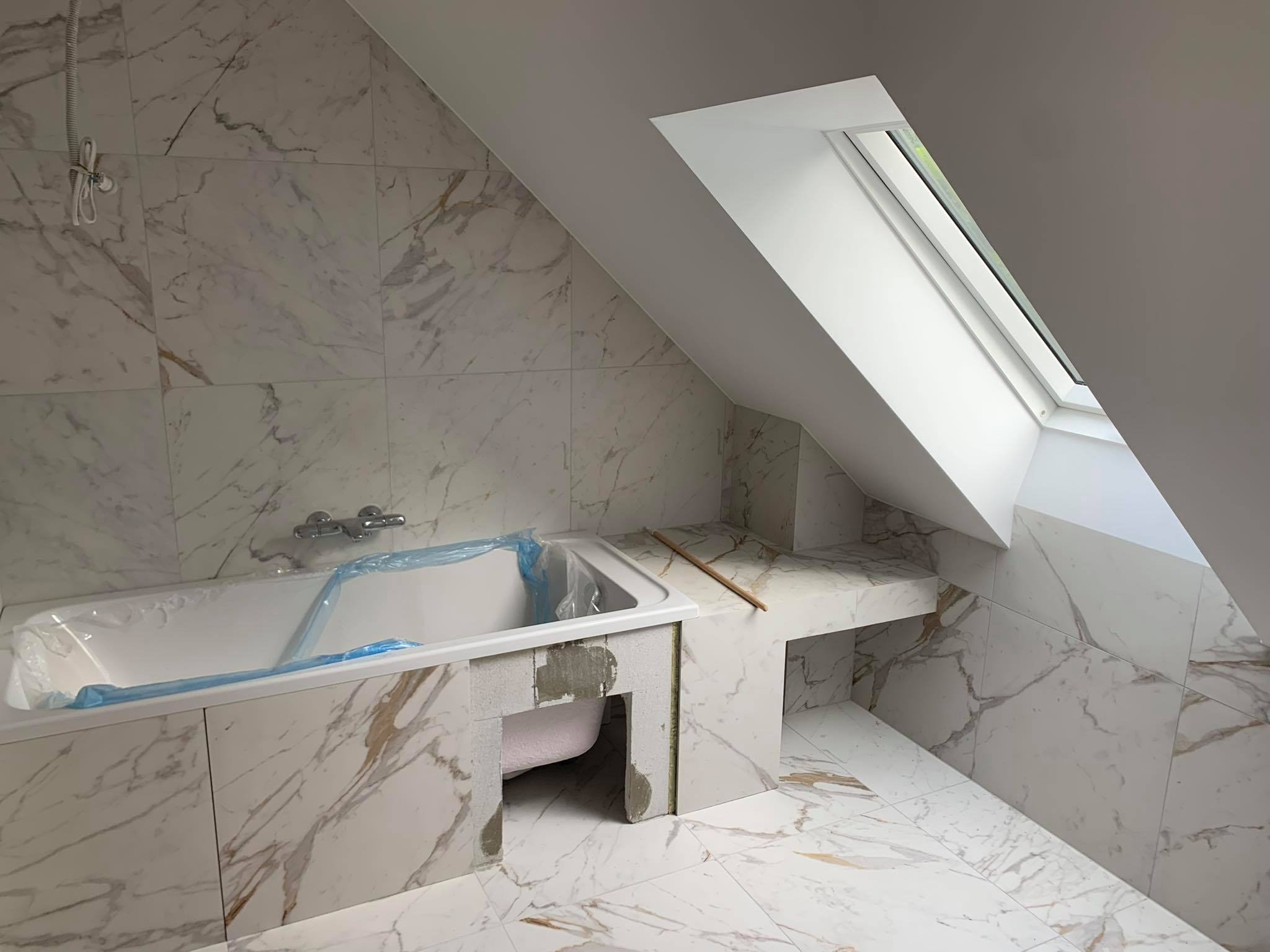 Remont łazienki na poddaszu: białe marmurowe płytki, zabudowana wanna z folią ochronną, okno dachowe, etap prac wykończeniowych.