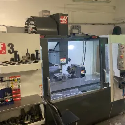 Wnętrze warsztatu z maszyną CNC Haas VF-3, widoczne narzędzia skrawające, panel sterowania z ekranem, obrabiany element w uchwycie, oświetlenie robocze i elementy wyposażenia stanowiska.