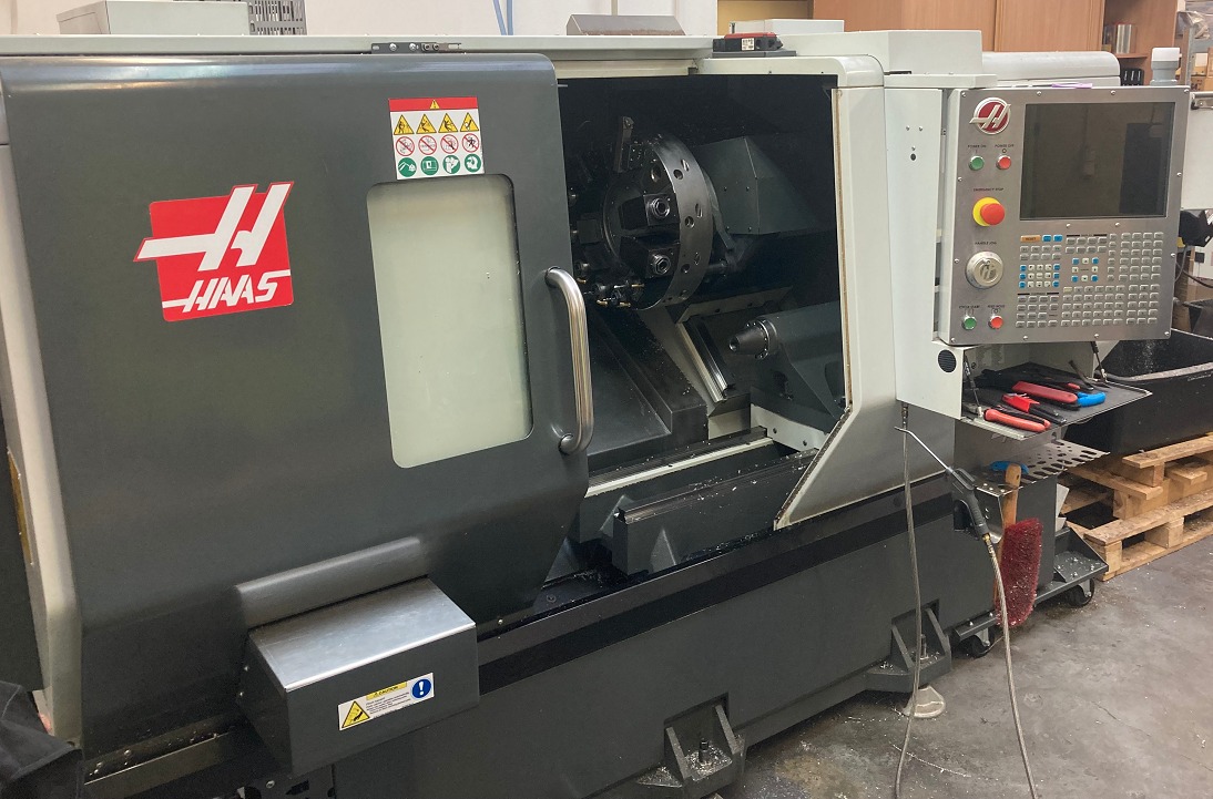 Otwarta obudowa nowoczesnej tokarki CNC marki Haas, widoczne wnętrze z uchwytem narzędziowym, panel sterowania z przyciskami i ekranem, oraz narzędzia na półce bocznej.